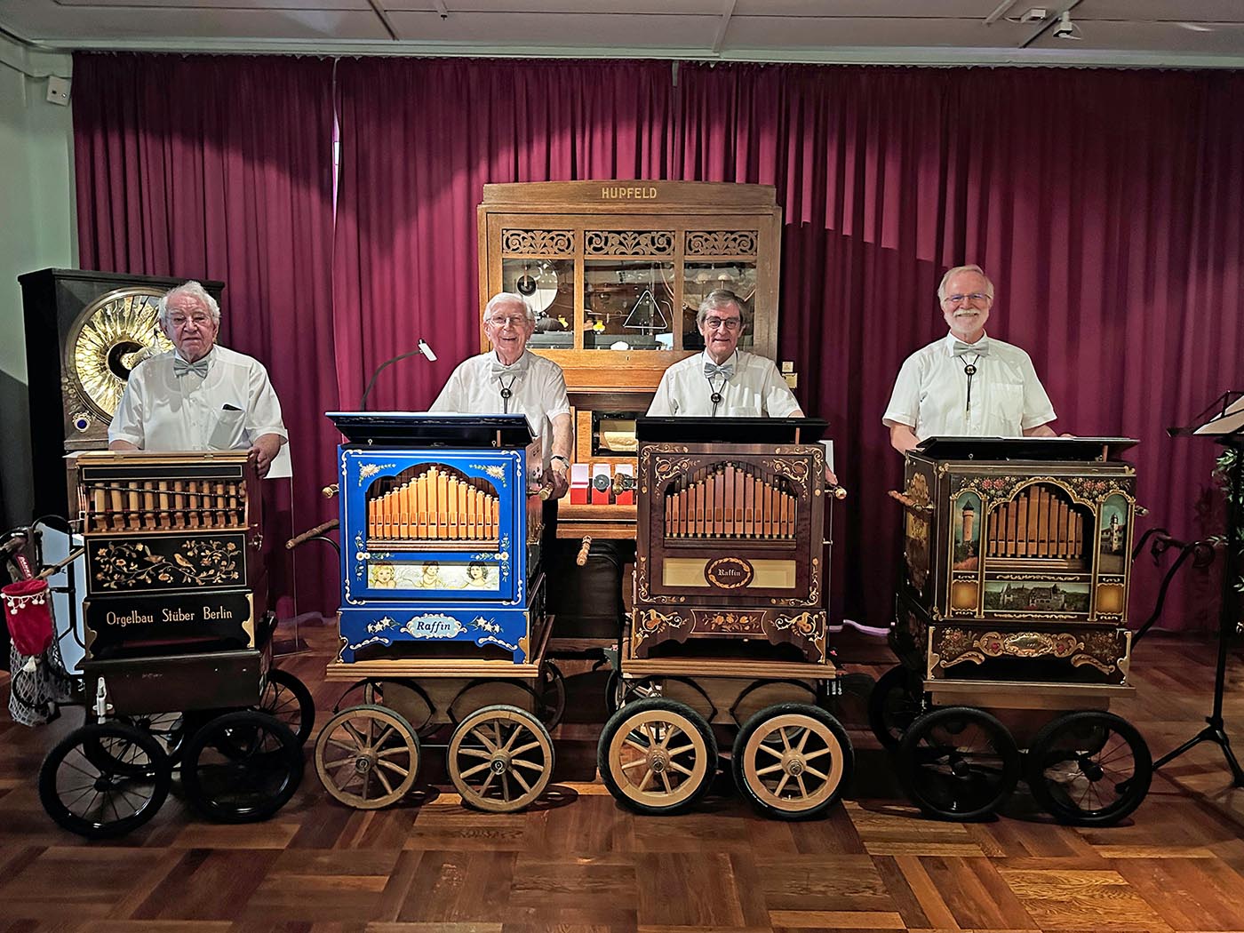 Vier Museums-Drehorgler mit historischen Drehorgeln auf einer Bühne vor rotem Vorhang.